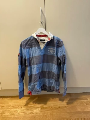Randig rugbytröja  - Säljer en långärmad rugbytröja med breda blå och grå ränder, vit krage och knappslå. Tröjan har broderad logga på bröstet och brittisk flagga på ärmen. Perfekt för en sportig och avslappnad stil.