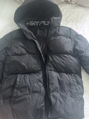 Svart pufferjacka från Tommy Hilfiger - Snygg svart pufferjacka från Tommy Hilfiger med huva och dragkedja. Jackan har logotyp på bröstet och ärmen samt stora quiltade partier som ger en cool look. Perfekt för kalla dagar och riktigt varm. Materialet är glansigt och vattenavvisande. liten defekt på högra fickan som ej syns vid användning.