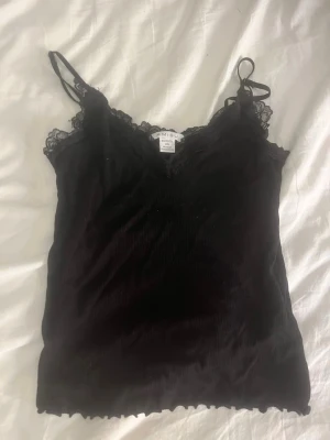 Svart linnetopp med spets från Amisu - Snygg svart linnetopp från Amisu med tunna axelband och spetsdetaljer vid urringningen. Toppen har v-ringad hals och är tillverkad i ett mjukt, ribbat material. Perfekt att styla med jeans eller kjol för en trendig look.
