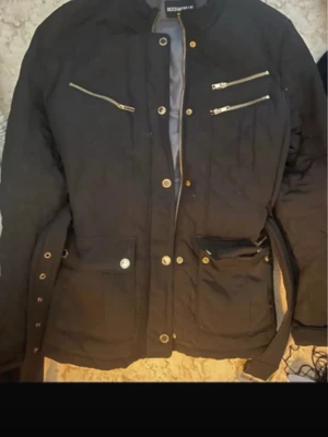 Svart jacka med dragkedjor från rockanblue  - Svart jacka från Boohoo med guldfärgade dragkedjor och knappar. Jackan har flera fickor, bälte i midjan och lång ärm. Perfekt för dig som gillar en stilren look med snygga detaljer.