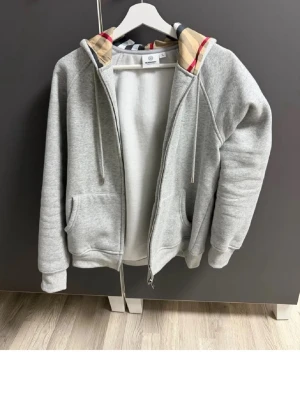 Grå zip hoodie från Burberry - Snygg grå hoodie från Burberry med dragkedja och klassiskt rutigt mönster i huvan. Hoodien har fickor framtill, ribbade muddar och justerbar dragsko i huvan. Perfekt för en chill och stilren look. Materialet är mjukt och bekvämt, troligen bomullsblandning. Storlek L men passar bäst till M