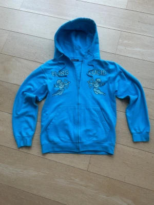 Blå hoodie med tryck från Bling Lucid - Snygg blå hoodie med dragkedja från Bling Lucid. Hoodien har huva och fickor framtill. Perfekt för dig som gillar streetwear och vill sticka ut lite extra. Tröjan är i toppen skick och alla pärlor är kvar. Tveka inte att höra av er vid frågor!!