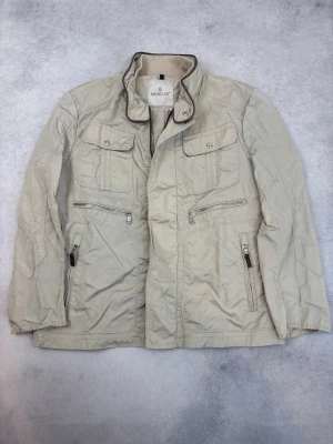 Beige jacka från Moncler med dragkedjor - Stilren beige jacka från Moncler med hög krage och flera fickor med dragkedjor och knappar. Jackan har lång ärm och en modern, något loose passform. Perfekt för dig som gillar klassisk men ändå trendig stil.