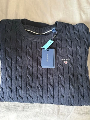 Mörkblå kabelstickad tröja GANT - Helt ny kabelstickad tröja från GANT i mörkblå färg med rund halsringning och ribbade muddar. Tröjan har GANT:s broderade logotyp med sköld på bröstet. Klassisk och tidlös design, perfekt för lager på lager. Materialet är mjukt och håller formen. Pris-499 kr. Helt ny! Inköpt för ca 1000 kr