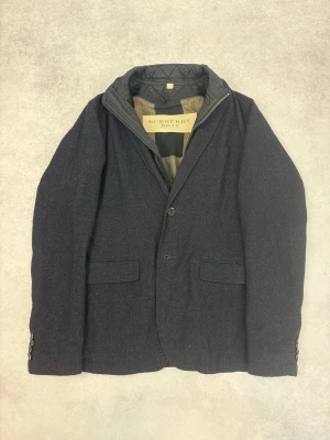 Mörkblå kavaj från Burberry Brit - Stilren mörkblå kavaj från Burberry Brit med tvåknäppning och klassisk passform. Kavajen har en quiltad innerkrage med dragkedja, två framfickor med lock och en bröstficka. Materialet är troligtvis ullblandning och insidan har Burberrys ikoniska rutmönster.