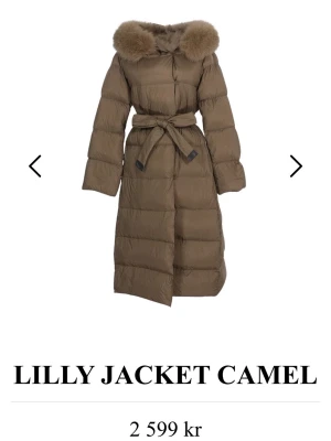 Lång camel dunjacka med pälskrage - Snygg lång dunjacka i camel från Lilly med stor fluffig pälskrage och knytskärp i midjan. Jackan har quiltad design och generös passform som håller dig varm under kalla vinterdagar. Perfekt för dig som vill ha både stil och värme.