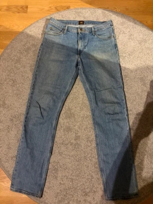 Ljusblå raka jeans från Lee - Säljer ett par ljusblå jeans från Lee med klassisk femficksmodell och raka ben. Jeansen har en snygg tvätt och Lee-logga på bakfickan. Tillverkade i denim med bälteshällor och dragkedjegylf. Perfekta för en avslappnad och trendig stil.