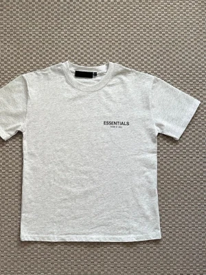 Essentials T-shirt Fear of God - Snygg ljusgrå T-shirt från Fear of God Essentials med svart logotyptryck fram och stort tryck på ryggen. Klassisk rund hals och rak passform. Tillverkad i mjuk bomull som känns skön mot huden. Perfekt för dig som gillar streetwear och enkel stil.