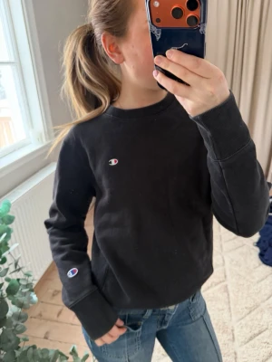 Svart sweatshirt från Champion - Svart sweatshirt från Champion med klassisk logga på bröstet och ärmen. Tröjan har rund halsringning och ribbade muddar vid ärmslut och midja. Perfekt för en avslappnad och sportig stil.