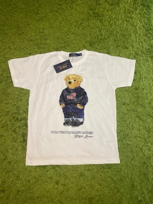 Vit t-shirt Polo Ralph Lauren björn - Vit t-shirt från Polo Ralph Lauren med ikoniskt björntryck på framsidan. Klassisk rund halsringning och korta ärmar. Tillverkad i mjuk bomull och har märkets logga tryckt under motivet. Perfekt statement-plagg för en trendig look.