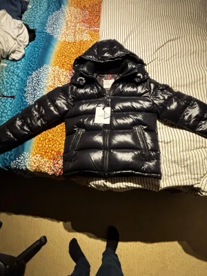 Svart pufferjacka från Moncler - Säljer en svart glansig pufferjacka från Moncler med huva och dragkedja framtill. Jackan har två snedställda fickor med dragkedja och ett ikoniskt serietidningsfoder på insidan. Perfekt för kalla dagar och riktigt snygg streetstil. Var inte blyg för o fråga ifall det är något. Priset går att diskutera!!