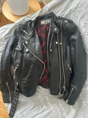 Svart skinnjacka från Jofama by Kenza  - Snygg svart skinnjacka från Jofama med coola detaljer som flera dragkedjor, axelklaffar och bälte i midjan. Insidan har rödsvart mönstrat foder. Klassisk bikerstil som ger en edgy look och passar perfekt till jeans eller kjol.