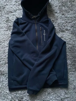 Mörkblå hoodie från Sail Racing - Snygg mörkblå hoodie från Sail Racing med dragkedja framtill och logga på bröstet. Hoodien har en stor huva med snörning och en praktisk bröstficka med dragkedja. Tillverkad i mjukt och bekvämt material, perfekt för chill dagar. Nypris 1500kr mitt pris 400kr pris går att diskutera vid snabb affär 