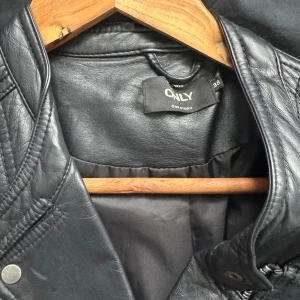 Svart skinnjacka från ONLY - Snygg svart skinnjacka från ONLY med asymmetrisk dragkedja, flera fickor med dragkedjor och coola sömdetaljer på axlarna. Klassisk bikerstil som ger en edgy look. Perfekt för dig som vill ha en tidlös jacka med attityd.