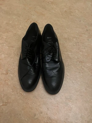 Svarta loafers från BOSS i läder - Snygga svarta loafers från BOSS i äkta läder med klassisk broguedetalj och snörning. Skorna har elegant perforerat mönster på tån och sidorna, samt en stilren och tidlös design som passar till många olika outfits.köpt för 5000 säljs för 1800