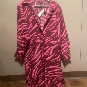 Zebramönstrad skjortklänning rosa - Zebramönstrad skjortklänning från H&M i stark rosa och röd ton. Klänningen har krage, knappar framtill och trekvartsärm. Perfekt för dig som vill sticka ut med ett djurmönster och en färgstark look.