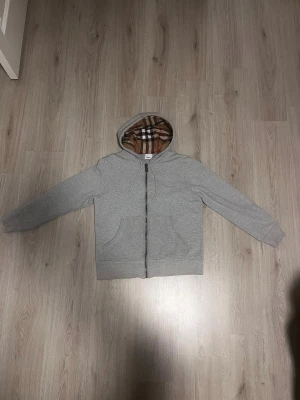 Grå hoodie med Burberry-mönstrad huva - Säljer en grå zip-hoodie från Burberry med klassiskt rutigt foder i huvan. Hoodien har dragkedja framtill, två fickor och justerbar dragsko i huvan. Perfekt för dig som gillar stilrena plagg med en twist. Materialet är mjukt och bekvämt bomullstyg. M Passar S också