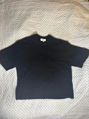 Svart stickad t-shirt från Gina  - Kortärmad svart stickad t-shirt från Arket med rund halsringning. Tröjan har en enkel och stilren design, perfekt för lager på lager eller som den är. Materialet ser mjukt och bekvämt ut, och passformen är något boxig.