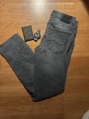 Grå jeans från Jack & Jones - Snygga grå jeans från Jack & Jones med klassisk femficksdesign och raka ben. Jeansen har en diskret tvättad look och svart patch med logga bak i midjan. Perfekta till en avslappnad stil och lätta att matcha med det mesta.