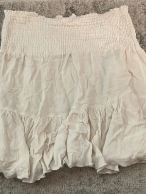 Vit volangkjol med smock - Supersöt kort vit kjol med bred smock i midjan och volangkant nertill. Kjolen har en luftig och flowy känsla, perfekt för varma dagar. Passar till både sneakers och sandaler för en chill look.