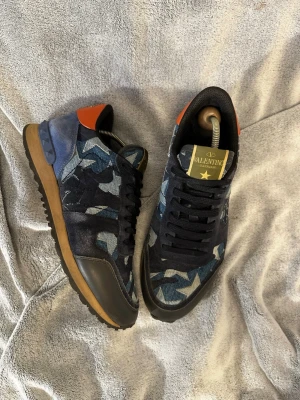 Valentino rockrunner camo sneakers i blått - Snygga sneakers från Valentino med blått och grått camomönster, svarta skosnören och detaljer i svart läder. Skorna har en brun sula och en orange detalj vid hälen. Perfekta för dig som vill sticka ut med stilrena och unika sneakers. Storlek 40 men passar 41