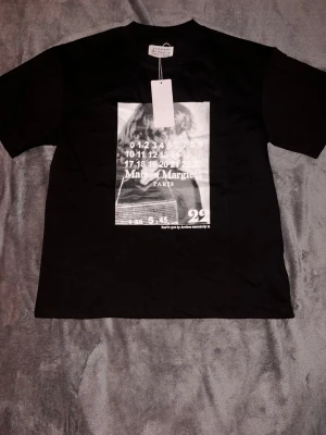 Maison Margiela  t-shirt - Svart t-shirt från Maison Margiela med stor grafisk print framtill i vitt och grått. Klassisk passform med rund hals och korta ärmar. Perfekt för dig som gillar streetstyle och vill sticka ut med en unik design.
