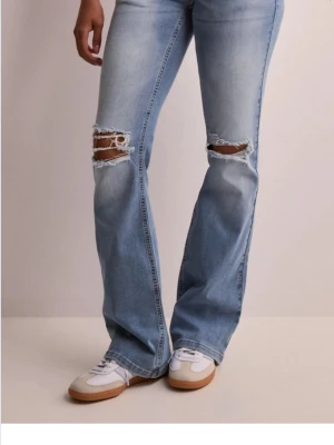 Ljusblå bootcut jeans med slitningar - Säljer ett par ljusblå bootcut jeans med slitna detaljer och hål vid knäna. Jeansen har en klassisk femficksmodell och är tillverkade i denim med lätt stretch. Perfekta för en avslappnad och trendig look. Från Nelly 