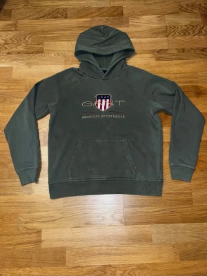 Grön hoodie från Gant med logga - Säljer en grön hoodie från Gant med broderad logga och texten 'American Sportswear' på bröstet. Hoodien har en stor ficka framtill och klassisk huva. Materialet är mjukt och skönt, perfekt för en avslappnad stil.