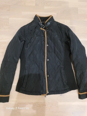 Quiltad jacka från Tindra - Quiltad svart jacka från Tindra med bruna detaljer längs kanterna och vid kragen. Jackan har långärmade ribbade sidor för extra komfort och en snygg spänndetalj vid halsen. Stängs med både dragkedja och knappar framtill. Perfekt för dig som gillar stilren design.