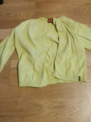 Gul kofta från Esprit - Ljusgul kofta från Esprit med enkel knappstängning framtill. Koftan har långa ärmar och ribbade muddar vid ärmslut och nederkant. Perfekt för att liva upp din outfit med en färgklick.