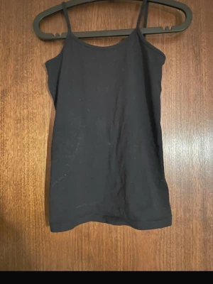 Svart baslinne med smala axelband - Enkel svart topp med smala axelband och rak passform. Perfekt att styla lager på lager eller bära som den är. Mjukt och stretchigt material för en bekväm känsla. Passar till många olika outfits.