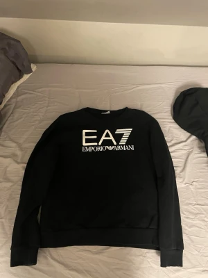 Svart sweatshirt EA7 Emporio Armani - Snygg svart sweatshirt från EA7 Emporio Armani med stort vitt logotryck på bröstet. Tröjan har rund halsringning och ribbade muddar vid ärmslut och nederkant. Perfekt för en trendig och avslappnad stil. Den är även i storlek S, använd fåtal gånger.