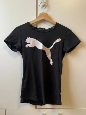 Svart Puma t-shirt med logga - Snygg svart t-shirt från Puma med den klassiska stora loggan i vitt framtill. T-shirten har rund halsringning och korta ärmar. Perfekt till jeans eller shorts för en sportig look.