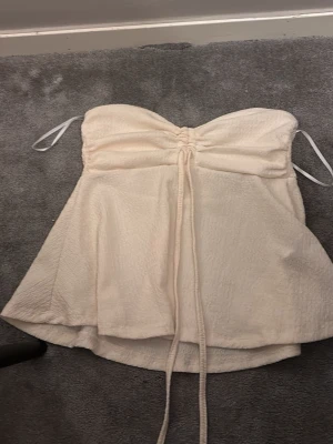 Krämvit bandeau topp med knytband - Säljer en krämvit bandeau topp med tunna axelband och snygg rynkning framtill. Toppen har en lätt utsvängd passform och långa knytband som ger en trendig look. Perfekt till sommarens outfits och enkel att styla med jeans eller kjol.