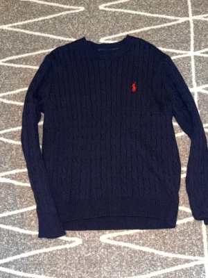 Mörkblå kabelstickad tröja Polo Ralph Lauren - Mörkblå kabelstickad tröja från Polo Ralph Lauren med rund halsringning och röd broderad logga på bröstet. Tröjan har långa ärmar och ribbade muddar vid ärmslut och nederkant. Perfekt för en stilren och klassisk look.