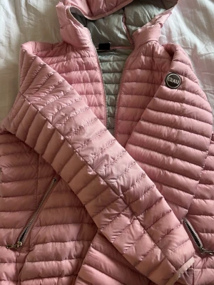 Ljusrosa dunjacka från Colmar - Snygg ljusrosa dunjacka från Colmar med huva och två dragkedjeförsedda fickor. Jackan har quiltad design och Colmar-logga på armen Perfekt för vinterdagar och dessutom nu till våren.