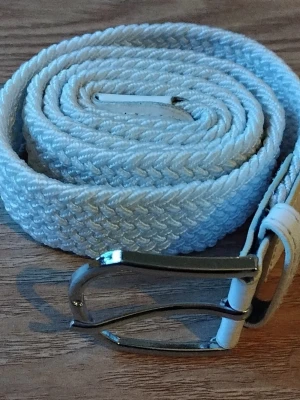 Vitt flätat bälte med silverfärgat spänne - Snyggt vitt flätat bälte med ett stilrent silverfärgat metallspänne. Bältet är gjort i ett mjukt syntetmaterial och har en klassisk design som passar till många olika outfits. Bältet är ungefär 107 cm långt.