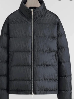 Svart pufferjacka från Dior - Säljer en svart pufferjacka från Dior med diskret monogrammönster över hela jackan. Jackan har hög krage, två sidofickor och dragkedja framtill. Perfekt för kalla dagar och riktigt snygg till streetwear-looken.