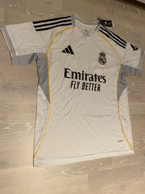Real Madrid matchtröja Adidas vit - Säljer en vit Real Madrid matchtröja från Adidas med svarta detaljer och gula linjer. Tröjan har klubbmärke på bröstet, Adidas-logga och sponsortryck 'Emirates Fly Better' framtill. Tillverkad i lätt och ventilerande material, perfekt för fotbollsträning.