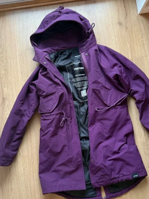 Lila regnjacka från Tretorn - Snygg lila regnjacka från Tretorn med huva, dragsko i midjan och stora fickor framtill. Jackan har broderad logga på ärmen och är tillverkad i ett vattenavvisande material. Perfekt för regniga dagar och har en längre passform för extra skydd.