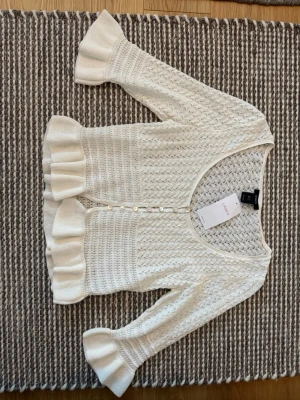 Vit stickad kofta med volangärm - Supersöt kräm-vit stickad kofta från Lindex med volangdetaljer vid ärmslut och nederkant. Koftan har knappar framtill, trekvartsärmar och ett fint kabelstickat mönster. Det är helt ny och aldrig använd! 