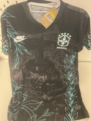 Nike Brasil matchtröja svart/blå - Snygg svart matchtröja från Nike med Brasil-logga och ikoniskt tryck av Kristusstatyn. Tröjan har blå detaljer, vita Nike- och Brasilmärken samt mönstrade ärmar. Tillverkad i lätt och ventilerande material, perfekt för fotboll eller träning.