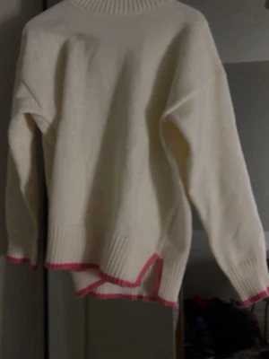 Vit stickad tröja med rosa detaljer - Säljer en oversized vit stickad tröja med ribbad polokrage och rosa kantdetaljer vid ärmslut och nederkant. Tröjan har en avslappnad passform och asymmetrisk nederkant som ger en cool touch. Perfekt för lager-på-lager-stil.