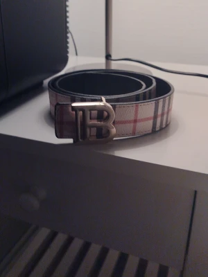 Rutig Burberry bälte med B-spänne - Snyggt skärp från Burberry med klassiskt rutigt mönster i beige, svart och rött. Skärpet har ett stort metallspänne med ett B framtill. Perfekt accessoar för att lyfta din outfit.