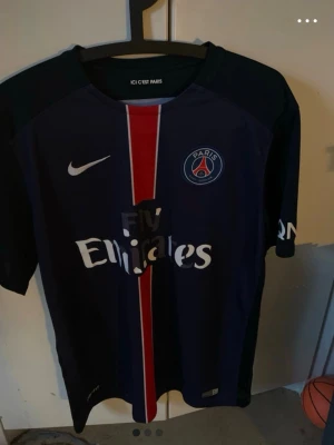 PSG matchtröja Nike Dri-FIT - Snygg PSG matchtröja från Nike med Dri-FIT material. Tröjan är mörkblå med röd och vit rand i mitten, Paris Saint-Germain klubbmärke på bröstet och Fly Emirates-tryck. Perfekt för fotbollsträning eller att bära till vardags.