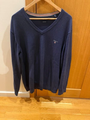 Mörkblå V-ringad tröja från Gant - Klassisk mörkblå V-ringad tröja från Gant med broderad logga på bröstet. Tröjan har långa ärmar och ribbade muddar vid ärmslut och nederkant. Perfekt för en stilren och avslappnad look.