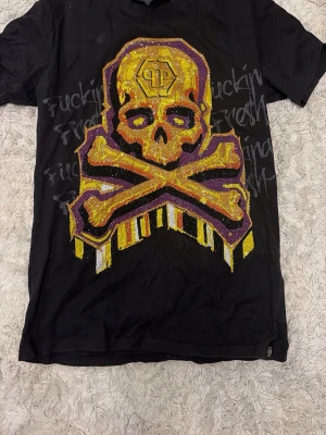 Svart Philipp Plein t-shirt med dödskalle - Säljer en svart t-shirt från Philipp Plein med stor dödskalle och korslagda ben i gult, lila och vitt framtill, dekorerad med glittrande stenar. Baksidan har lila graffiti-tryck och gul text. Materialet är mjuk bomull och modellen är rak.