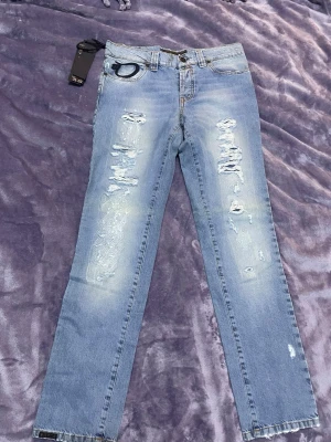 Blå slitna jeans med raka ben - Säljer ett par ljusblå jeans från D&G med raka ben och slitningar på framsidan. Jeansen har klassiska fem fickor, en D&G-logga på bakfickan och råa kanter längst ner. Materialet är denim och de har en cool, trendig look.