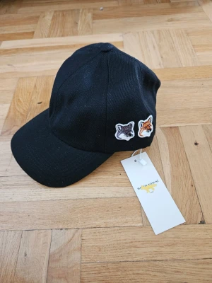 Svart keps Maison Kitsuné - Svart keps från Maison Kitsuné med två broderade rävmärken framtill och justerbart spänne bak. Kepsen har Maison Kitsuné-loggan broderad på sidan och är tillverkad i ett slitstarkt tyg. Perfekt accessoar för en trendig streetstyle.