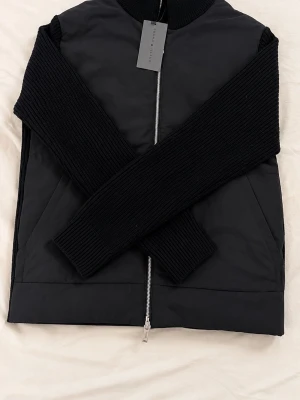 Svart cardigan från brand Island  - Säljer en superfin stickad svart cardigan/ hybridjacka från Brand Island. Modellen har en vadderad framsida som skyddar mot vind, medan ärmarna och ryggen är i en kraftig, ribbstickad kvalitet! Säljer pågrund av att jag aldrig använder den. Hör gärna av dig vid frågor eller funderingar😋